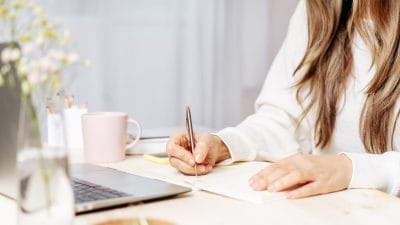 Person writing an obituary at a table with a laptop and flowers nearby.