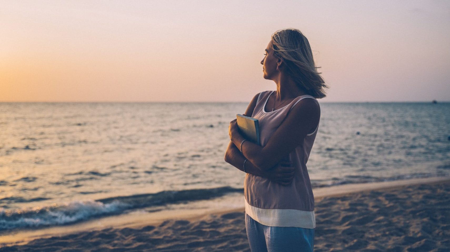 A woman stands on a beach at sunset, holding a book and gazing out over the ocean in a reflective moment.